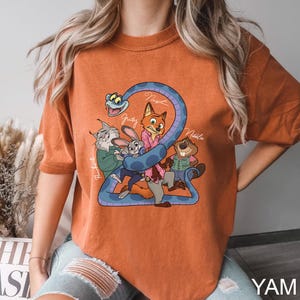 K&ouml;nnte beinhalten: Gebrannt orangefarbenes T-Shirt mit einer Cartoon-Grafik von Filmfiguren. Das Design umfasst eine blaue Schlange, die sich um die Zahl 2 windet, mit Charakternamen wie Judy und Nick. Das Shirt hat eine lockere Passform.