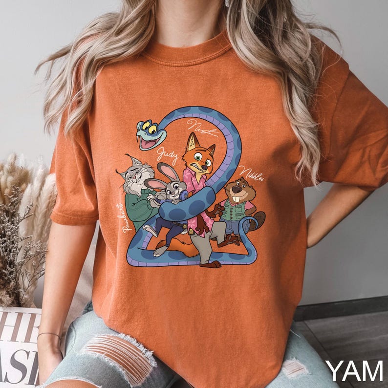 K&ouml;nnte beinhalten: Gebrannt orangefarbenes T-Shirt mit einer Cartoon-Grafik von Filmfiguren. Das Design umfasst eine blaue Schlange, die sich um die Zahl 2 windet, mit Charakternamen wie Judy und Nick. Das Shirt hat eine lockere Passform.