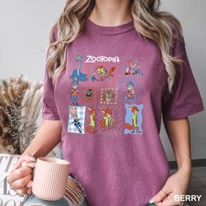 May include: Berry-colored t-shirt featuring a colorful graphic of characters from the animated film "Zootopia." The design includes the film's title and various character illustrations within decorative frames. The shirt is a comfortable, casual style.
