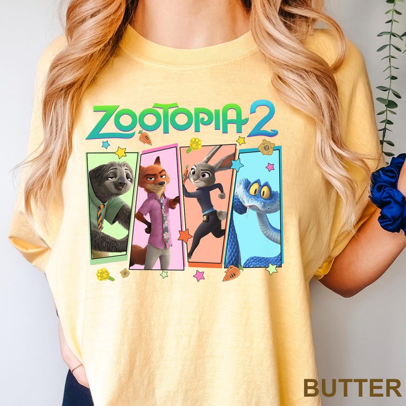 K&ouml;nnte beinhalten: Hellgelbes T-Shirt mit dem Schriftzug "ZOOTOPIA 2" &uuml;ber vier Cartoon-Charakter-Panels. Die Panels zeigen ein Faultier, einen Fuchs, ein Kaninchen und eine Schlange. Das Wort "BUTTER" ist unten rechts aufgedruckt.