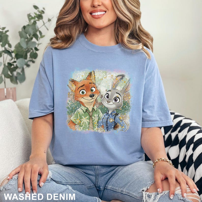 May include: A light blue t-shirt featuring a colorful graphic of the Zootopia characters Nick Wilde and Judy Hopps. The shirt has the words "WASHED DENIM" printed at the bottom.