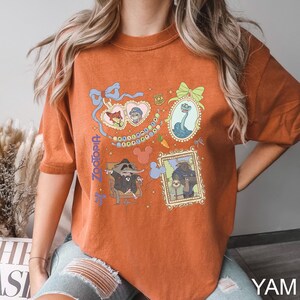 Puede incluir: Camiseta naranja quemado con ilustraciones enmarcadas de personajes de la pel&iacute;cula animada Zootopia. La camiseta incluye el t&iacute;tulo de la pel&iacute;cula en una fuente estilizada. Las ilustraciones est&aacute;n en marcos ornamentados con un estilo de dibujos animados.