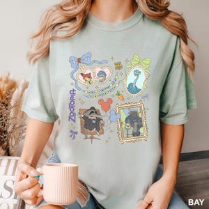Puede incluir: Camiseta verde claro con ilustraciones coloridas de personajes de la pel&iacute;cula "Zootr&oacute;polis". El dise&ntilde;o incluye retratos enmarcados, corazones y la palabra "Zootopia" en fuente vertical. La camiseta es de estilo casual y c&oacute;modo.