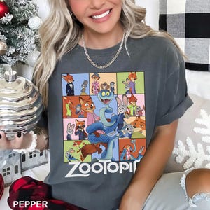 Puede incluir: Camiseta gris oscuro con un gr&aacute;fico colorido de personajes de la pel&iacute;cula animada "Zootopia". El dise&ntilde;o incluye una serpiente azul y varios personajes de animales en un patr&oacute;n de cuadr&iacute;cula. La palabra "ZOOTOPIA" est&aacute; impresa en blanco.