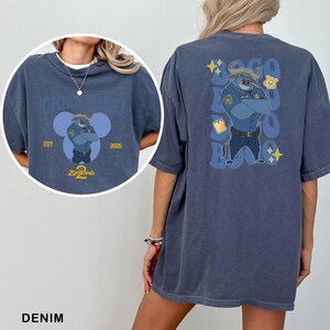 May include: Denim blue t-shirt featuring a cartoon police officer from Zootopia. The front has a circular design with the text "Zootopia 2" and "EST 2025". The back displays the officer with text and star graphics. A casual, oversized fit.