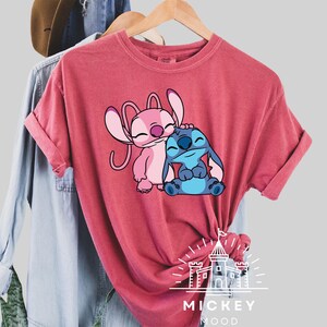 May include: A red t-shirt with a graphic of Stitch and Angel from Lilo & Stitch. The graphic shows Stitch hugging Angel. The shirt has the text "Mickey Mood" on the bottom.