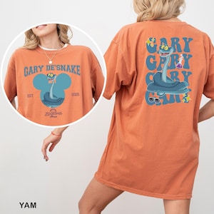 May include: Orange t-shirt with a cartoon snake design. The front features the text "GARY DE'SNAKE" above a blue snake illustration. The back of the shirt repeats the text and snake design. The shirt includes the text "Zootopia 2" and the year "2025".