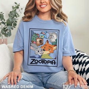 May include: Light blue t-shirt featuring a graphic from the animated film Zootopia. The graphic includes characters Judy Hopps, Nick Wilde, and others, with the film's title in bold black letters. The shirt is labeled "Washed Denim".