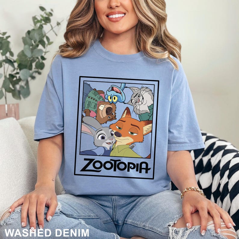 May include: Light blue t-shirt featuring a graphic from the animated film Zootopia. The graphic includes characters Judy Hopps, Nick Wilde, and others, with the film's title in bold black letters. The shirt is labeled "Washed Denim".