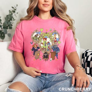 May include: A pink t-shirt featuring a cartoon graphic of animal characters in ornate frames. The design includes a central heart-shaped element with a bow and stars. The word "CRUNCHBERRY" is printed at the bottom.