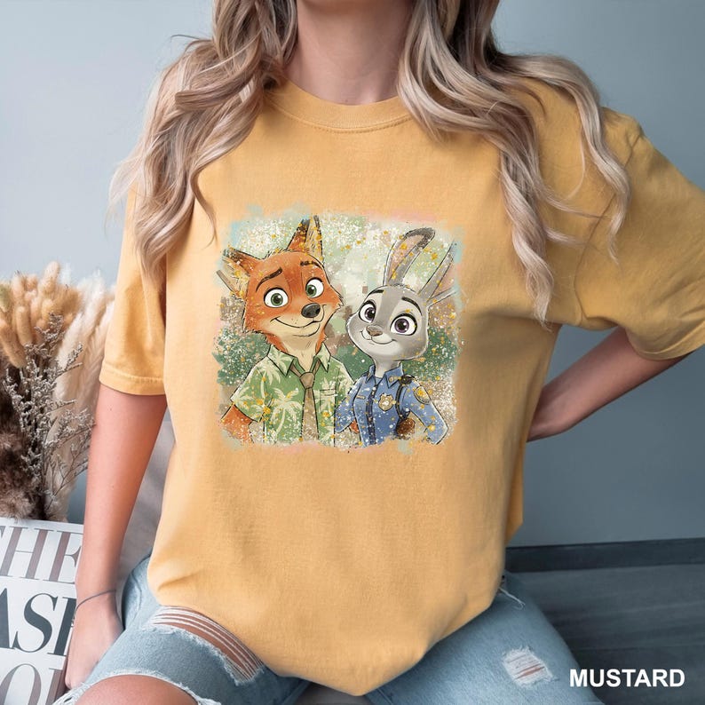 May include: Mustard-colored t-shirt featuring a graphic of Nick Wilde and Judy Hopps from Zootopia. Nick is a fox wearing a green shirt, and Judy is a rabbit in a police uniform. The shirt has a distressed, vintage look.