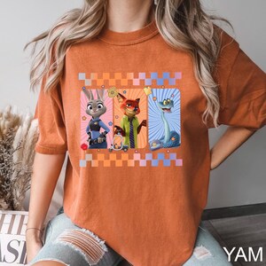 Puede incluir: Camiseta naranja quemado con un gr&aacute;fico de tres personajes de dibujos animados: un conejo, un zorro y una serpiente. Los personajes est&aacute;n sobre un fondo a cuadros. La camiseta tiene mangas cortas y un corte holgado. El texto "YAM" es visible en la esquina inferior derecha.