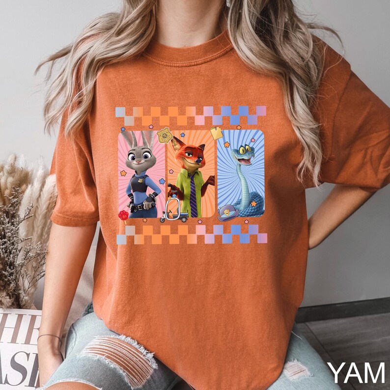 Puede incluir: Camiseta naranja quemado con un gr&aacute;fico de tres personajes de dibujos animados: un conejo, un zorro y una serpiente. Los personajes est&aacute;n sobre un fondo a cuadros. La camiseta tiene mangas cortas y un corte holgado. El texto "YAM" es visible en la esquina inferior derecha.