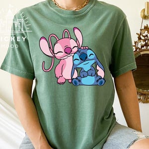 May include: A green t-shirt with a cartoon image of Stitch and Angel from the Disney movie Lilo & Stitch. Stitch is blue and Angel is pink.