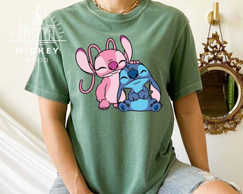 May include: A green t-shirt with a cartoon image of Stitch and Angel from the Disney movie Lilo & Stitch. Stitch is blue and Angel is pink.
