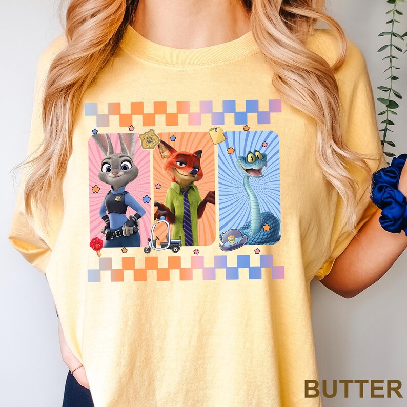 Puede incluir: Camiseta amarilla con tres personajes de Zootopia: Judy Hopps, Nick Wilde y una serpiente. Cada personaje est&aacute; en un marco rectangular con un color de fondo diferente. La palabra "BUTTER" est&aacute; impresa en la parte inferior derecha.