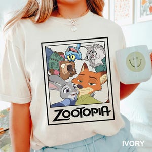 May include: Ivory-colored t-shirt featuring a framed graphic of characters from the animated film "Zootopia." The graphic includes Judy Hopps, Nick Wilde, and other characters. The word "Zootopia" is printed in bold, black letters below the characters.