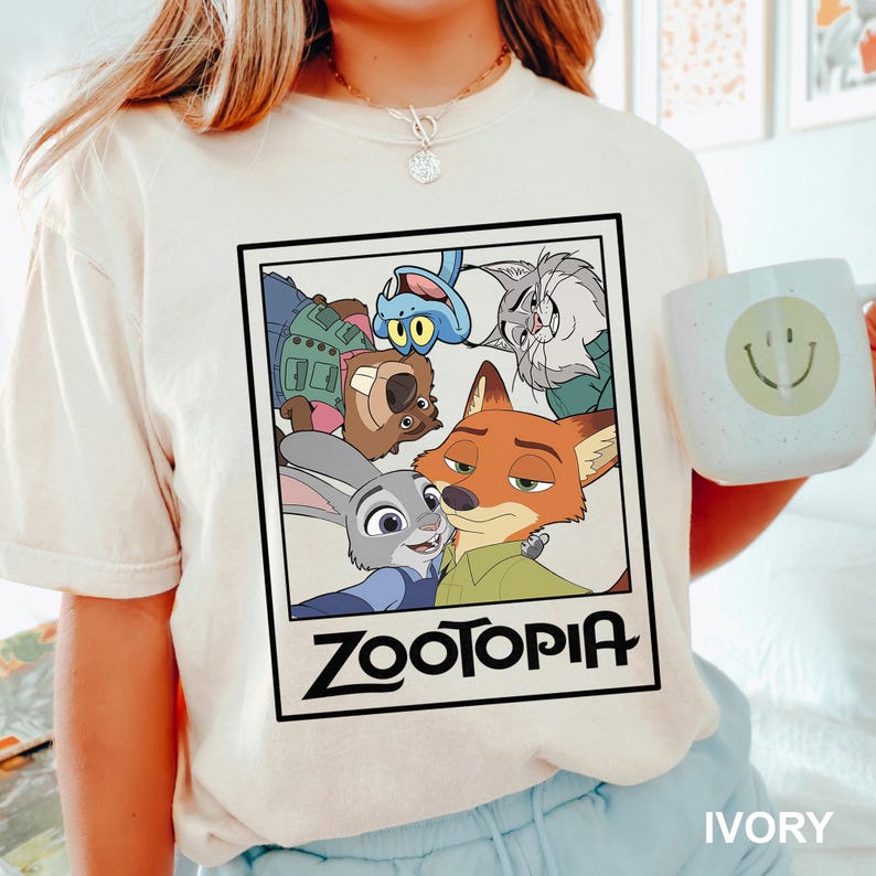 May include: Ivory-colored t-shirt featuring a framed graphic of characters from the animated film "Zootopia." The graphic includes Judy Hopps, Nick Wilde, and other characters. The word "Zootopia" is printed in bold, black letters below the characters.