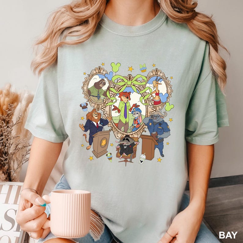 May include: A light green t-shirt featuring a colorful cartoon graphic of various animal characters in a heart-shaped frame. The graphic includes a fox, a rabbit, a lion, and other animals. The word "BAY" is printed in white at the bottom right.