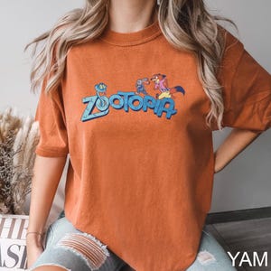 Op de afbeelding: Oranje-bruin T-shirt met het woord "Zootopia" in blauw en stripfiguren uit de film. Het shirt heeft korte mouwen en een ronde hals. Het T-shirt is gemaakt van een zacht materiaal.