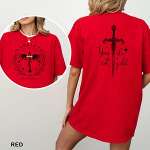 Może przedstawiać: Czerwona koszulka z czarną grafiką z przodu i z tyłu. Grafika z przodu zawiera tekst "BASGIATH WAR COLLEGE" i "DRAGON RIDER" z motywem miecza i smoka. Grafika z tyłu przedstawia motyw miecza i słowa "You do not yield."