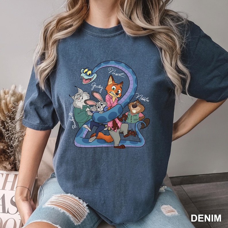 K&ouml;nnte beinhalten: Denimblaues T-Shirt mit einer Cartoon-Grafik von Filmfiguren. Die Grafik zeigt einen Fuchs, ein Kaninchen, einen Biber und eine Schlange. Die Namen der Charaktere sind dar&uuml;ber geschrieben. Das Wort "DENIM" ist unten rechts aufgedruckt.