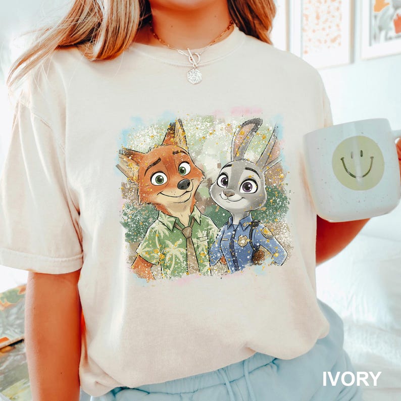 May include: Ivory-colored t-shirt featuring a colorful illustration of Nick Wilde and Judy Hopps from Zootopia. Nick is wearing a Hawaiian shirt, and Judy is in a police uniform. A white mug with a smiley face is also visible.