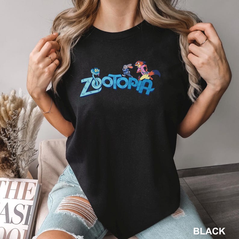 Op de afbeelding: Zwart T-shirt met ronde hals met het woord "Zootopia" in blauw en kleurrijke cartoonfiguren. Het shirt heeft een klassieke pasvorm en is gemaakt van zacht materiaal. Het woord "BLACK" staat rechtsonder gedrukt.
