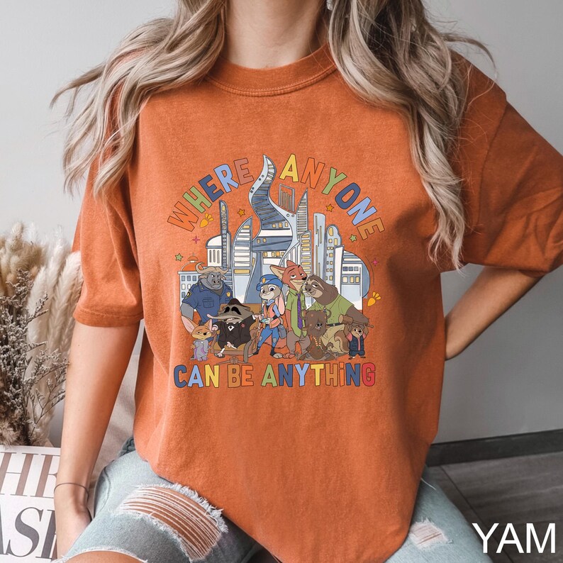 May include: Burnt orange t-shirt with a colourful graphic of Zootopia characters and the text "WHERE ANYONE CAN BE ANYTHING". The design includes a cityscape and various animal characters from the animated film.