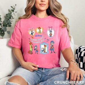 May include: A pink t-shirt with a design from the Disney film Zootopia. The graphic includes cartoon images of Judy Hopps and Nick Wilde, the word "Zootopia", and the phrase "welcome to Zootopia".