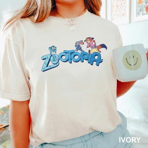 Op de afbeelding: Ivoor T-shirt met het woord "Zootopia" in blauw en stripfiguren. Een witte mok met een smiley wordt in de afbeelding vastgehouden. Het shirt heeft een casual stijl en een comfortabele pasvorm.
