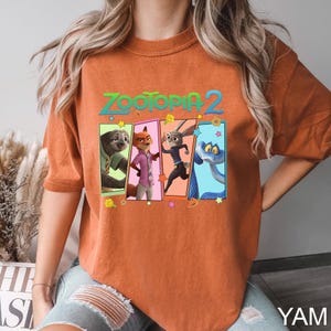 K&ouml;nnte beinhalten: Gebrannt orangefarbenes T-Shirt mit dem Text "ZOOTOPIA 2" &uuml;ber vier Charakterportr&auml;ts. Die Charaktere sind ein Faultier, ein Fuchs, ein Kaninchen und eine Schlange. Das Shirt ist ein bequemer, l&auml;ssiger Stil.