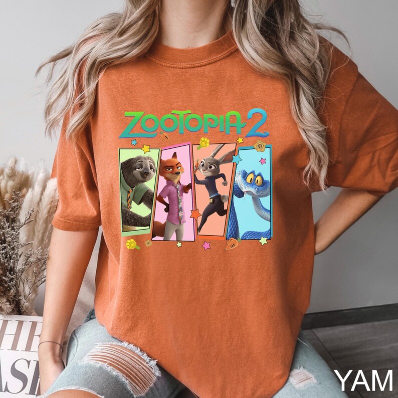 K&ouml;nnte beinhalten: Gebrannt orangefarbenes T-Shirt mit dem Text "ZOOTOPIA 2" &uuml;ber vier Charakterportr&auml;ts. Die Charaktere sind ein Faultier, ein Fuchs, ein Kaninchen und eine Schlange. Das Shirt ist ein bequemer, l&auml;ssiger Stil.