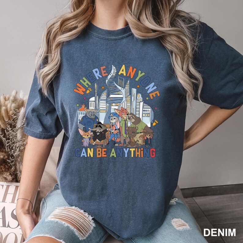 May include: A dark blue t-shirt with a vibrant graphic of cartoon characters and a city skyline. The text reads "WHERE ANYONE CAN BE ANYTHING" in a multi-coloured font.