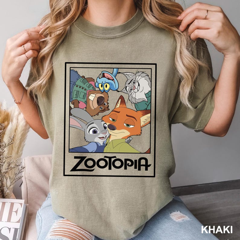 May include: Khaki-colored t-shirt featuring a framed graphic of characters from the animated film "Zootopia." The graphic includes Judy Hopps, Nick Wilde, and other characters. The word "Zootopia" is printed in a bold, black font.
