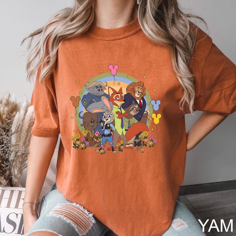May include: Burnt orange t-shirt featuring a colorful cartoon graphic of animal characters. The design includes a rainbow, flowers, and several characters in suits and uniforms. The shirt has a relaxed fit and short sleeves.