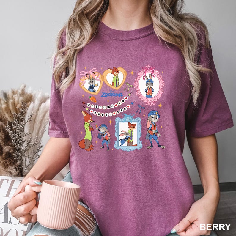 May include: Berry-coloured t-shirt featuring cartoon characters from the movie Zootopia. The design includes images of a fox and a rabbit in various frames and poses, along with the word "Zootopia". The shirt has a crew neck and short sleeves.