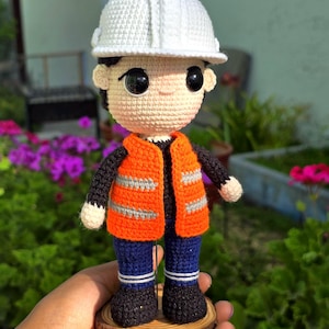 Puede incluir: Una muñeca de ganchillo de un trabajador de la construcción con casco blanco, chaleco naranja de alta visibilidad con dos franjas reflectantes plateadas, camisa negra de manga larga, pantalones azul oscuro y botas negras.