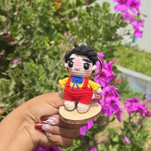 May include: A handmade crochet doll keychain. The doll has black hair, a yellow shirt, red overalls, and a blue bow tie. It is attached to a wooden base and a metal keyring. The background features green foliage and pink flowers.