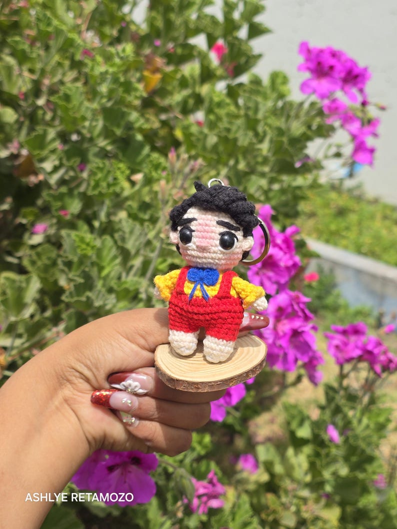 May include: A handmade crochet doll keychain. The doll has black hair, a yellow shirt, red overalls, and a blue bow tie. It is attached to a wooden base and a metal keyring. The background features green foliage and pink flowers.