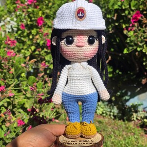 May include: A crocheted doll featuring long black hair, a white hard hat with a small emblem, a white long-sleeved shirt, blue pants, and yellow shoes. The doll stands on a circular wooden base engraved with the text ING. KIARA A. ESTRELLA ZEA ROQUE.