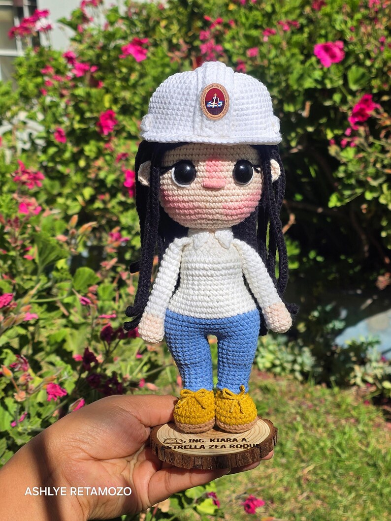 May include: A crocheted doll featuring long black hair, a white hard hat with a small emblem, a white long-sleeved shirt, blue pants, and yellow shoes. The doll stands on a circular wooden base engraved with the text ING. KIARA A. ESTRELLA ZEA ROQUE.