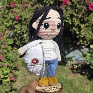 May include: A crocheted doll with black yarn hair, wearing a white long-sleeve shirt, blue pants, and yellow shoes. The doll holds a white hard hat with a circular emblem and stands on a wooden base engraved with "ING. KIARA A. ESTRELLA ZEA ROQU".