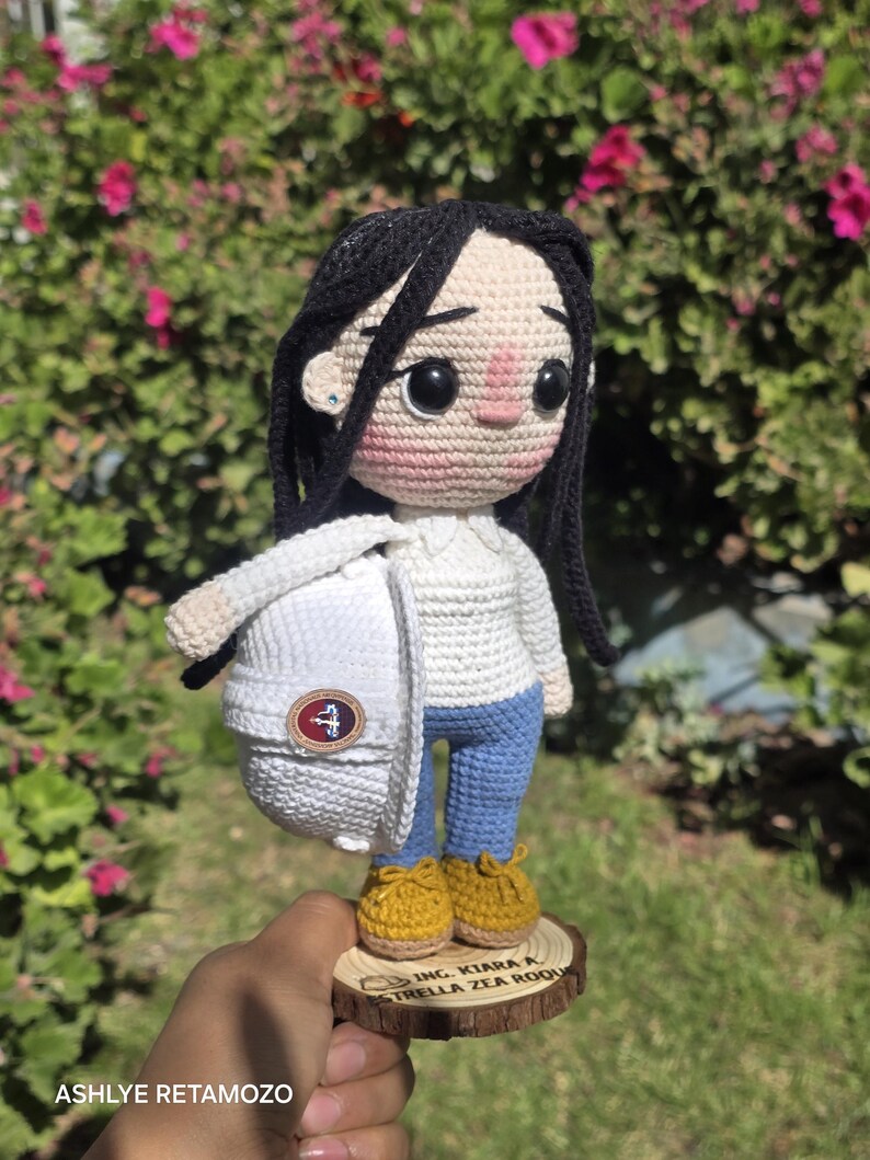 May include: A crocheted doll with black yarn hair, wearing a white long-sleeve shirt, blue pants, and yellow shoes. The doll holds a white hard hat with a circular emblem and stands on a wooden base engraved with "ING. KIARA A. ESTRELLA ZEA ROQU".