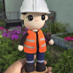 May include: A crocheted doll wearing a white hard hat, orange safety vest, blue pants, and black shoes. The doll is standing on a wooden base.