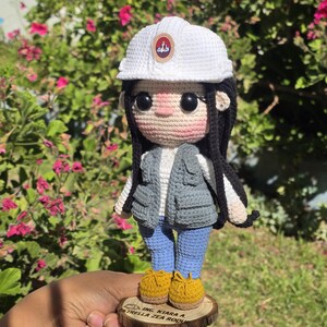 May include: A crochet doll wearing a white hard hat with a circular emblem, a grey utility vest over a white shirt, blue pants, and yellow shoes. The doll has long black hair and stands on a circular wooden base with the text ING. KIARA A. ESTRELLA ZEA ROQUE.