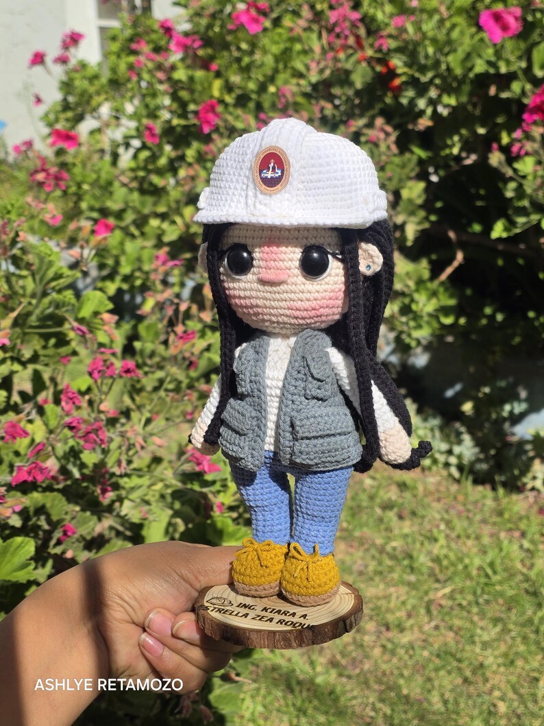 May include: A crochet doll wearing a white hard hat with a circular emblem, a grey utility vest over a white shirt, blue pants, and yellow shoes. The doll has long black hair and stands on a circular wooden base with the text ING. KIARA A. ESTRELLA ZEA ROQUE.