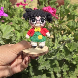 May include: A handmade crochet doll of a girl with black hair in pigtails, wearing glasses, a red jacket, a green dress, and a yellow bow. The doll is mounted on a small, round wooden base. The doll is held in front of a background of green foliage and pink flowers.