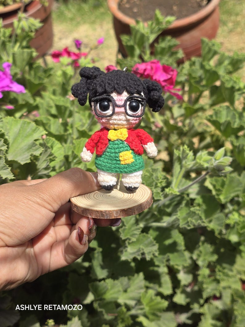 May include: A handmade crochet doll of a girl with black hair in pigtails, wearing glasses, a red jacket, a green dress, and a yellow bow. The doll is mounted on a small, round wooden base. The doll is held in front of a background of green foliage and pink flowers.