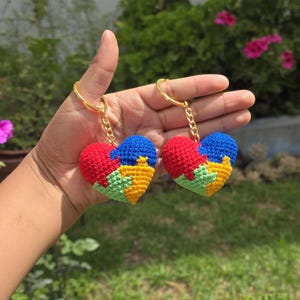 Puede incluir: Dos llaveros de corazón tejidos a crochet con un diseño de pieza de rompecabezas. Los llaveros son rojos, azules, verdes y amarillos. Los llaveros están unidos a cadenas doradas.